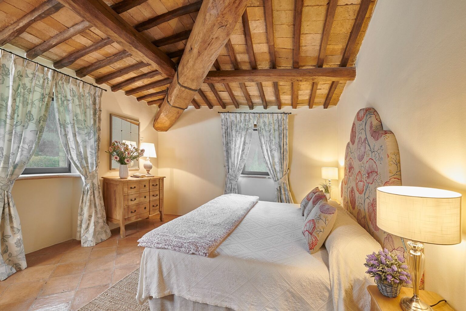 Cozy bedroom featuring wooden beamed ceiling, floral curtains, a comfortable bed with decorative pillows, a bedside lamp, and a wooden dresser with a vase of flowers, creating a warm and inviting atmosphere. | Only on SopranoVillas