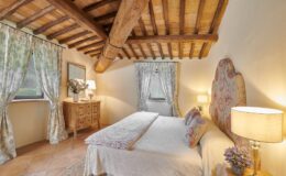 Cozy bedroom featuring wooden beamed ceiling, floral curtains, a comfortable bed with decorative pillows, a bedside lamp, and a wooden dresser with a vase of flowers, creating a warm and inviting atmosphere.