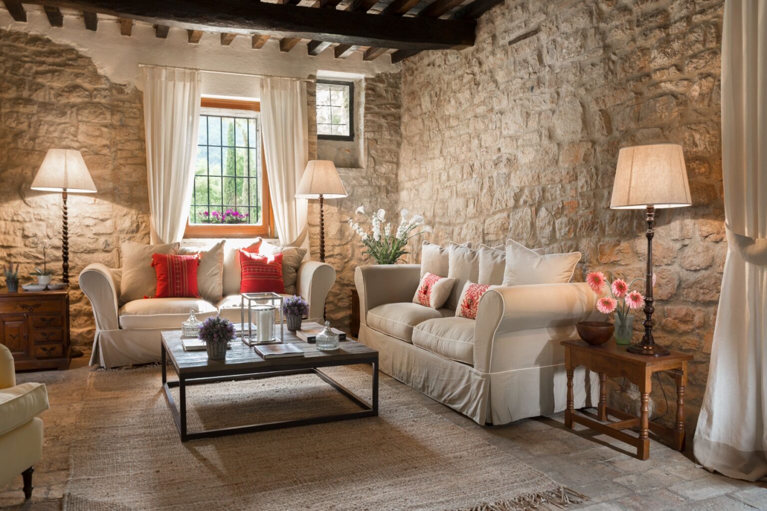 Cozy living room featuring natural stone walls, two comfortable sofas with decorative pillows, wooden furniture, and stylish lighting. The space is enhanced by a large window with sheer curtains, allowing natural light and a view of greenery outside. A rustic coffee table holds decorative plants and candles, creating a warm and inviting atmosphere. | Only on SopranoVillas