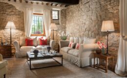 Cozy living room featuring natural stone walls, two comfortable sofas with decorative pillows, wooden furniture, and stylish lighting. The space is enhanced by a large window with sheer curtains, allowing natural light and a view of greenery outside. A rustic coffee table holds decorative plants and candles, creating a warm and inviting atmosphere.