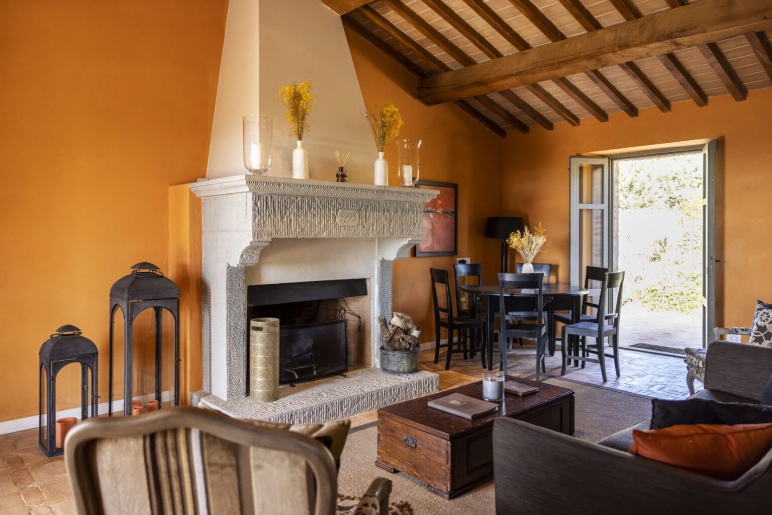 Cozy living room with an orange wall, featuring a stone fireplace, wooden beams, a round dining table, and stylish decor. The space includes black lanterns, a wooden coffee table, and an open door leading to an outdoor area. | Only on SopranoVillas