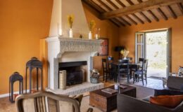 Cozy living room with an orange wall, featuring a stone fireplace, wooden beams, a round dining table, and stylish decor. The space includes black lanterns, a wooden coffee table, and an open door leading to an outdoor area.