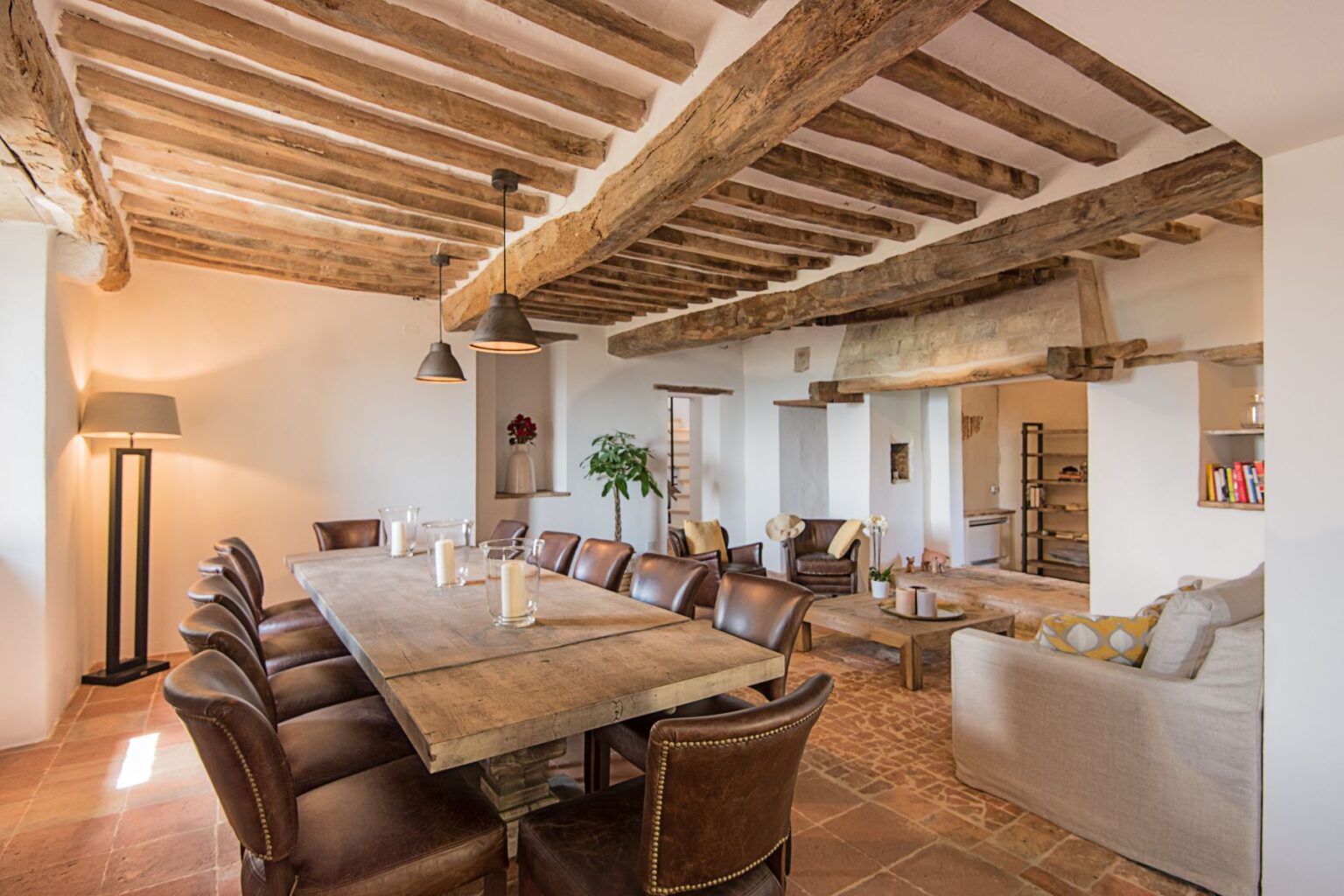 Spacious dining area featuring a large wooden table surrounded by leather chairs, complemented by rustic wooden beams and pendant lighting. The room includes a cozy seating area with a sofa and armchairs, adding to the inviting atmosphere. Natural light illuminates the space, highlighting the warm tones of the terracotta flooring. | Only on SopranoVillas