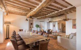 Spacious dining area featuring a large wooden table surrounded by leather chairs, complemented by rustic wooden beams and pendant lighting. The room includes a cozy seating area with a sofa and armchairs, adding to the inviting atmosphere. Natural light illuminates the space, highlighting the warm tones of the terracotta flooring.