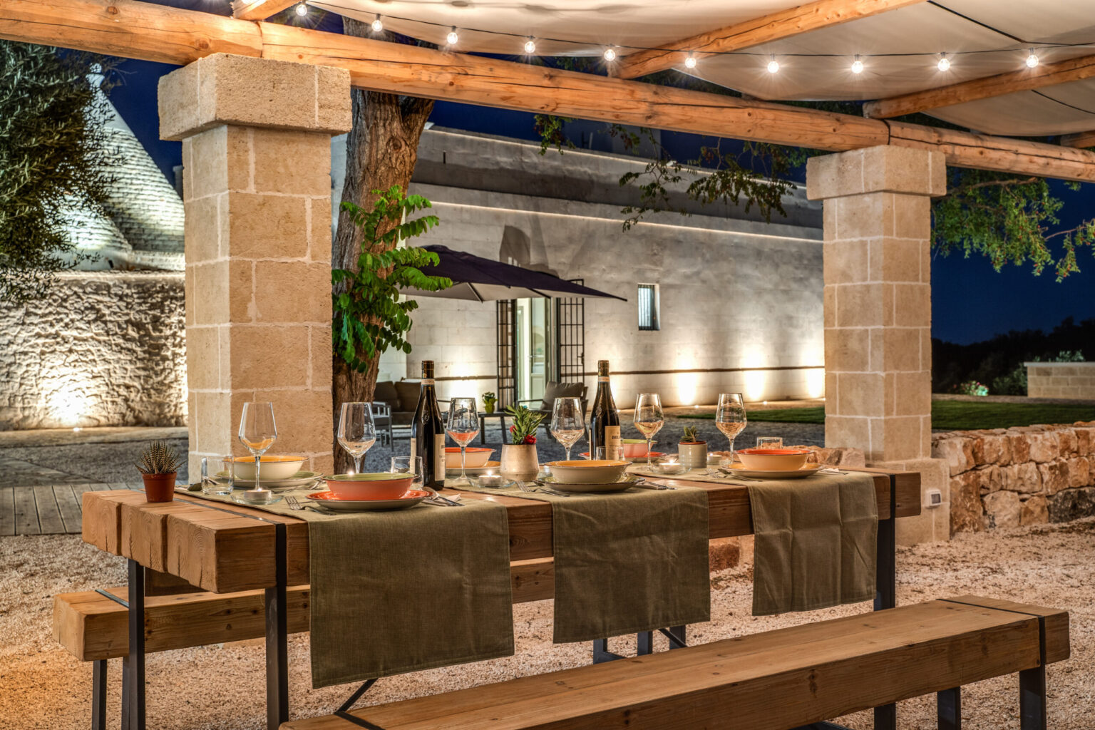Outdoor dining setup at night with a beautifully arranged table featuring elegant tableware, surrounded by warm lighting and a rustic stone building in the background, creating a cozy and inviting atmosphere. | Only on SopranoVillas
