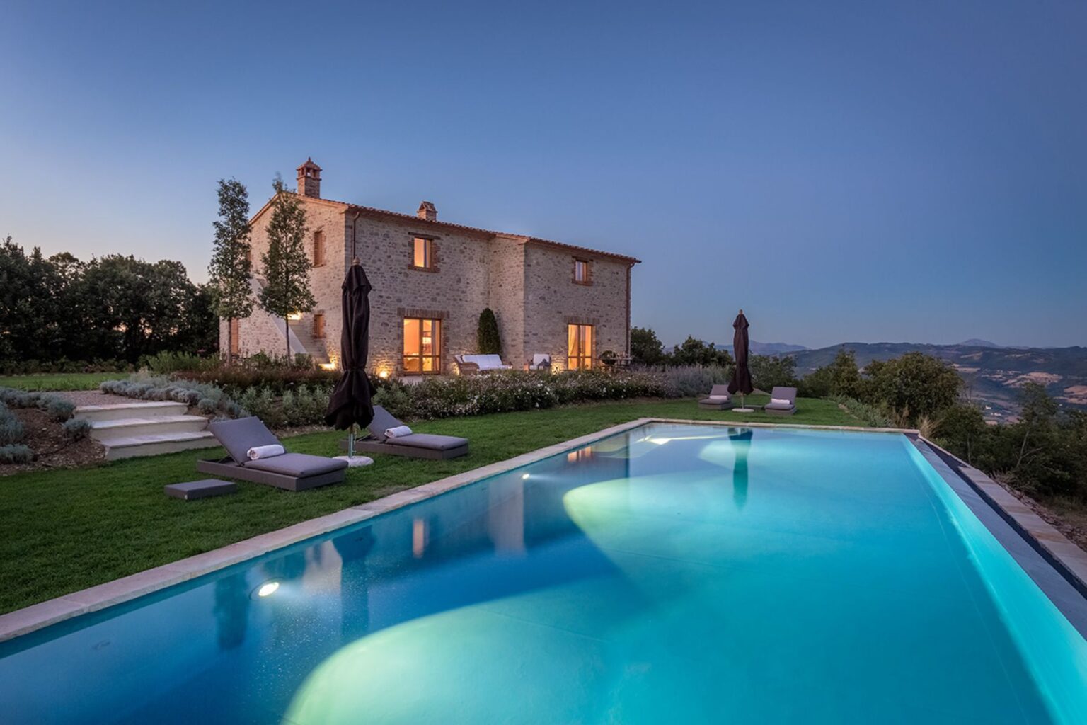 Scenic view of a rustic stone villa at dusk, featuring a sparkling swimming pool surrounded by lush greenery and lounge chairs, set against a backdrop of rolling hills and a clear sky. | Only on SopranoVillas