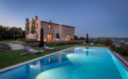 Scenic view of a rustic stone villa at dusk, featuring a sparkling swimming pool surrounded by lush greenery and lounge chairs, set against a backdrop of rolling hills and a clear sky.
