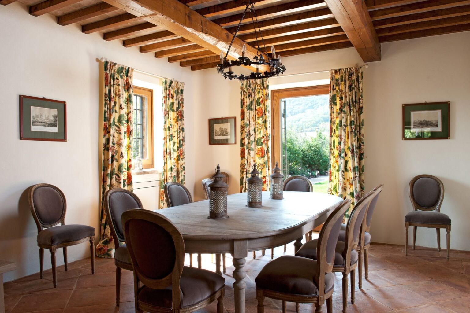 Cozy dining room featuring a rustic wooden table surrounded by elegant chairs, adorned with floral curtains and warm, natural light streaming through the windows. The room has exposed wooden beams and decorative lanterns, creating a welcoming atmosphere. | Only on SopranoVillas