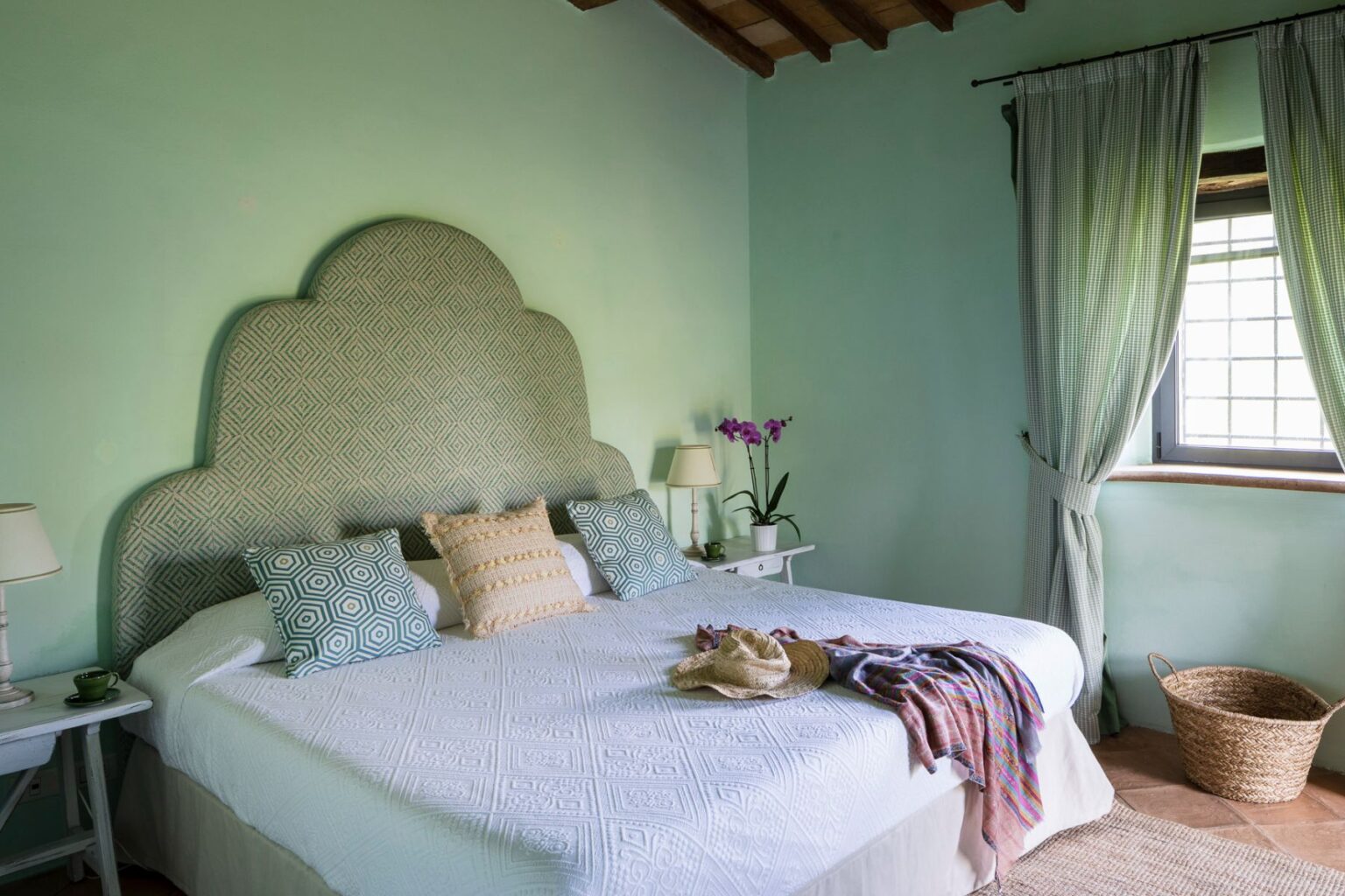 Cozy bedroom featuring a light green wall, a patterned headboard, and a neatly made bed with decorative pillows. Natural light streams in through a window adorned with soft curtains, complemented by a bedside lamp and a potted orchid. A woven basket adds a rustic touch to the serene atmosphere. | Only on SopranoVillas