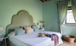 Cozy bedroom featuring a light green wall, a patterned headboard, and a neatly made bed with decorative pillows. Natural light streams in through a window adorned with soft curtains, complemented by a bedside lamp and a potted orchid. A woven basket adds a rustic touch to the serene atmosphere.