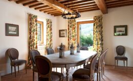 Cozy dining room featuring a rustic wooden table surrounded by elegant chairs, adorned with floral curtains and warm, natural light streaming through the windows. The room has exposed wooden beams and decorative lanterns, creating a welcoming atmosphere.