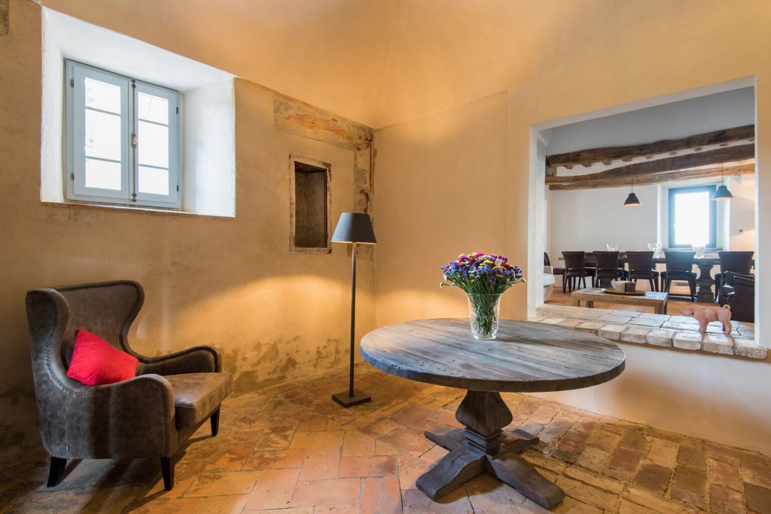 Cozy interior of a rustic room featuring a wooden table, a comfortable armchair with a red cushion, a vase of flowers, and a window that allows natural light. A glimpse of a dining area is visible through an open archway, enhancing the inviting atmosphere. | Only on SopranoVillas