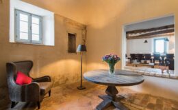 Cozy interior of a rustic room featuring a wooden table, a comfortable armchair with a red cushion, a vase of flowers, and a window that allows natural light. A glimpse of a dining area is visible through an open archway, enhancing the inviting atmosphere.