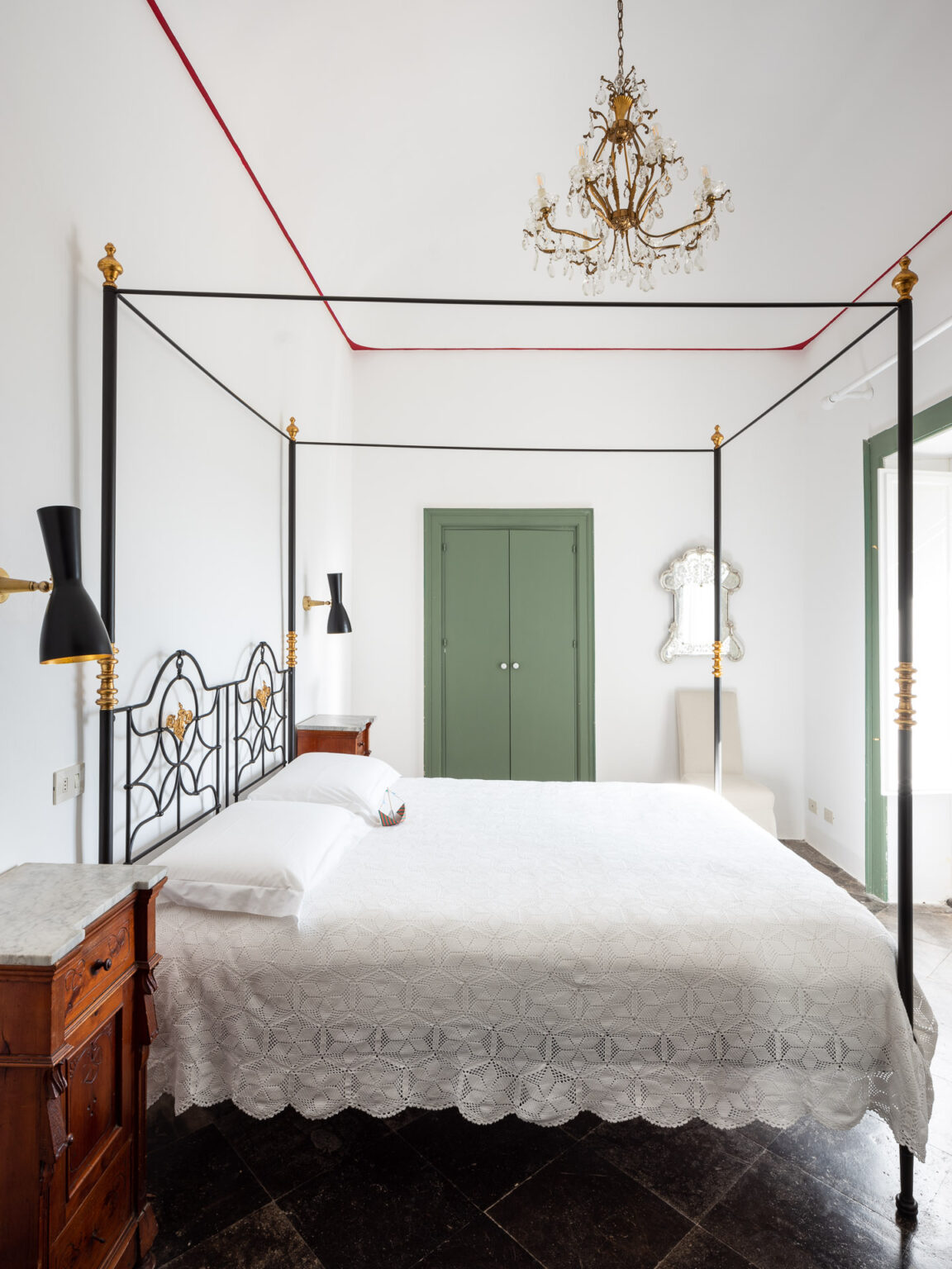 Elegant bedroom featuring a black four-poster bed with gold accents, a white lace bedspread, and a vintage chandelier. The room has marble-topped nightstands, a green door, and a mirror, all set against a bright white backdrop that highlights the stylish decor. | Only on SopranoVillas