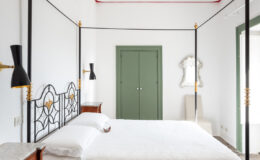 Elegant bedroom featuring a black four-poster bed with gold accents, a white lace bedspread, and a vintage chandelier. The room has marble-topped nightstands, a green door, and a mirror, all set against a bright white backdrop that highlights the stylish decor.