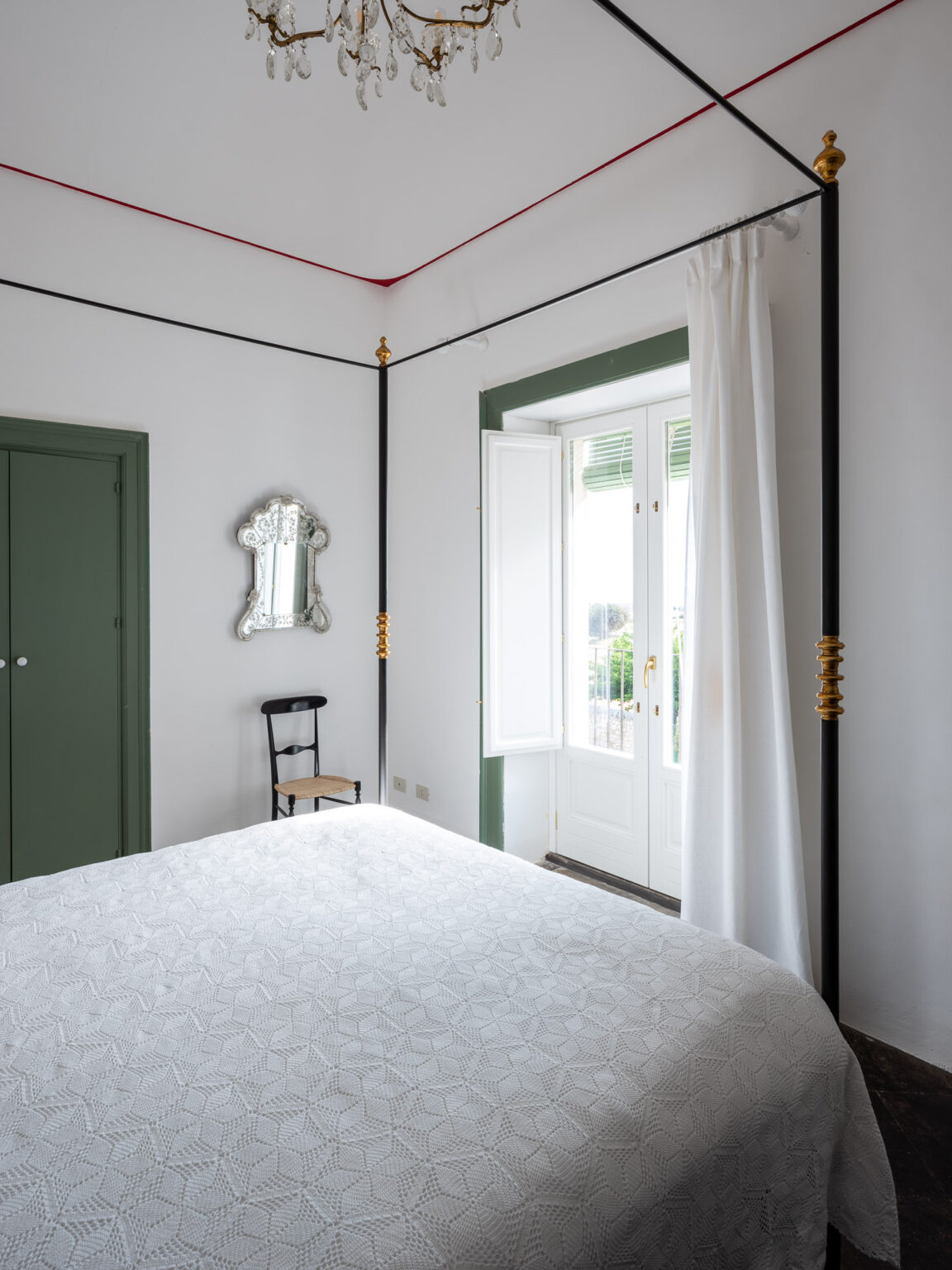 Bright and airy bedroom featuring a white quilted bedspread, a stylish chandelier, and elegant decor. The room includes a vintage mirror, a black chair, and large windows with white curtains, allowing natural light to illuminate the space. Green accents on the door and window frame add a touch of color to the minimalist design. | Only on SopranoVillas