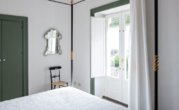 Bright and airy bedroom featuring a white quilted bedspread, a stylish chandelier, and elegant decor. The room includes a vintage mirror, a black chair, and large windows with white curtains, allowing natural light to illuminate the space. Green accents on the door and window frame add a touch of color to the minimalist design.