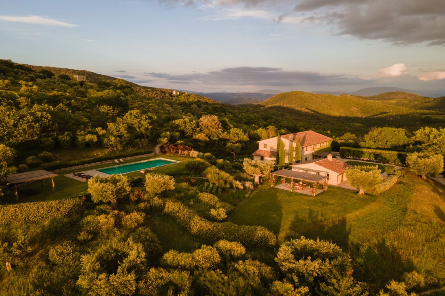 Aerial view of a picturesque countryside villa surrounded by lush greenery, featuring a swimming pool and landscaped gardens, set against rolling hills under a sunset sky. Ideal for relaxation and nature retreats. | Only on SopranoVillas