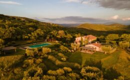 Aerial view of a picturesque countryside villa surrounded by lush greenery, featuring a swimming pool and landscaped gardens, set against rolling hills under a sunset sky. Ideal for relaxation and nature retreats.