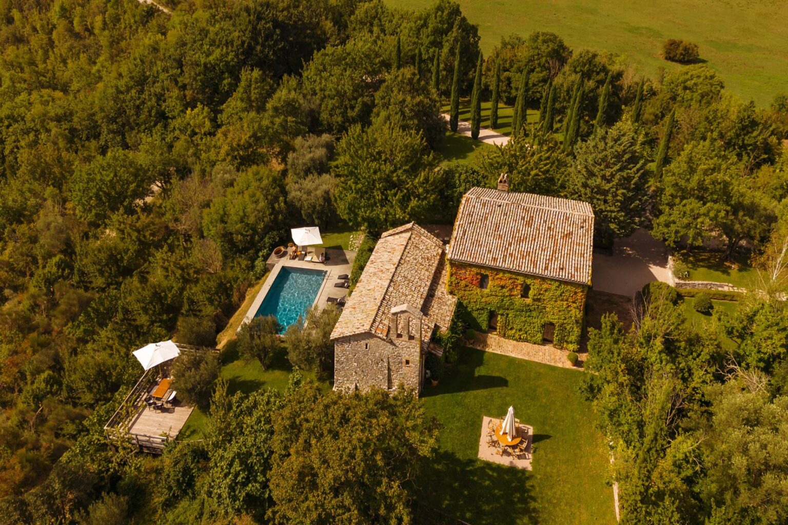 Aerial view of a charming countryside villa featuring a stone house, a swimming pool, and landscaped gardens surrounded by lush greenery. The image showcases outdoor seating areas and sun loungers, perfect for relaxation and enjoying nature. | Only on SopranoVillas