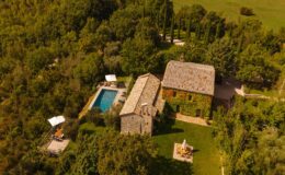 Aerial view of a charming countryside villa featuring a stone house, a swimming pool, and landscaped gardens surrounded by lush greenery. The image showcases outdoor seating areas and sun loungers, perfect for relaxation and enjoying nature.