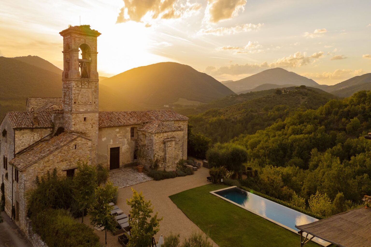 Historic stone building with a bell tower set against a sunset backdrop in a mountainous landscape, featuring a swimming pool and lush greenery in the foreground. Perfect for travelers seeking picturesque views and serene accommodations. | Only on SopranoVillas