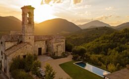 Historic stone building with a bell tower set against a sunset backdrop in a mountainous landscape, featuring a swimming pool and lush greenery in the foreground. Perfect for travelers seeking picturesque views and serene accommodations.