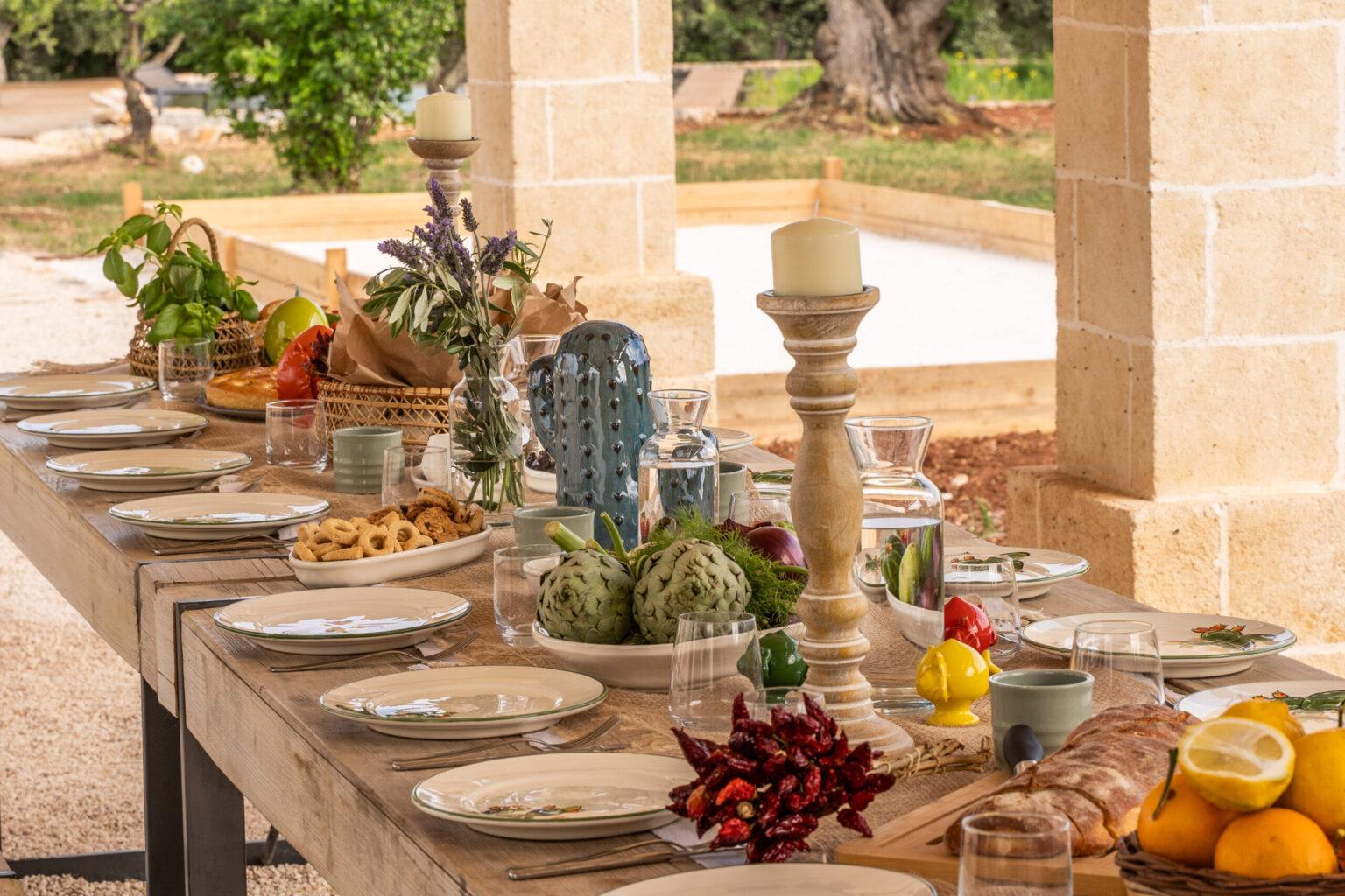 Beautiful outdoor dining setup featuring a long wooden table adorned with elegant tableware, fresh fruits, decorative candles, and vibrant centerpieces, set against a scenic garden backdrop. Perfect for outdoor gatherings and celebrations. | Only on SopranoVillas