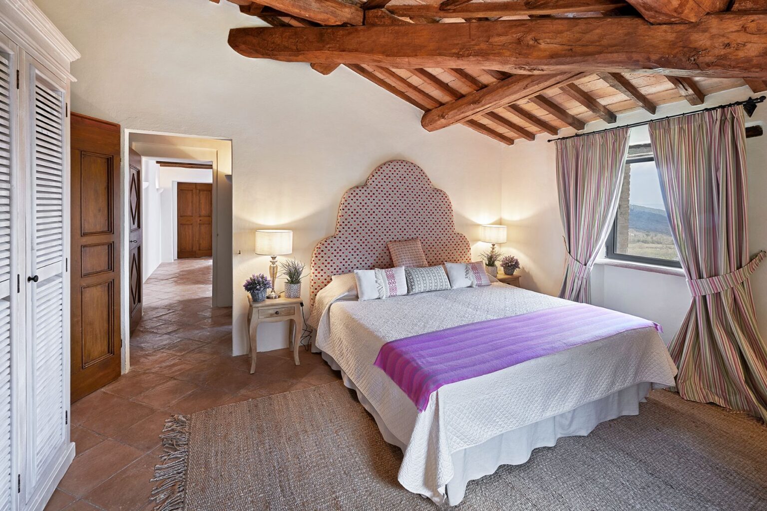 Cozy bedroom featuring a large bed with a purple throw blanket, decorative pillows, wooden ceiling beams, and stylish curtains. The room includes a bedside table with lamps and potted plants, creating a warm and inviting atmosphere. A doorway leads to a hallway with wooden doors, showcasing a blend of rustic and modern design elements. | Only on SopranoVillas