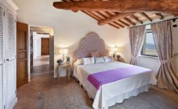 Cozy bedroom featuring a large bed with a purple throw blanket, decorative pillows, wooden ceiling beams, and stylish curtains. The room includes a bedside table with lamps and potted plants, creating a warm and inviting atmosphere. A doorway leads to a hallway with wooden doors, showcasing a blend of rustic and modern design elements.