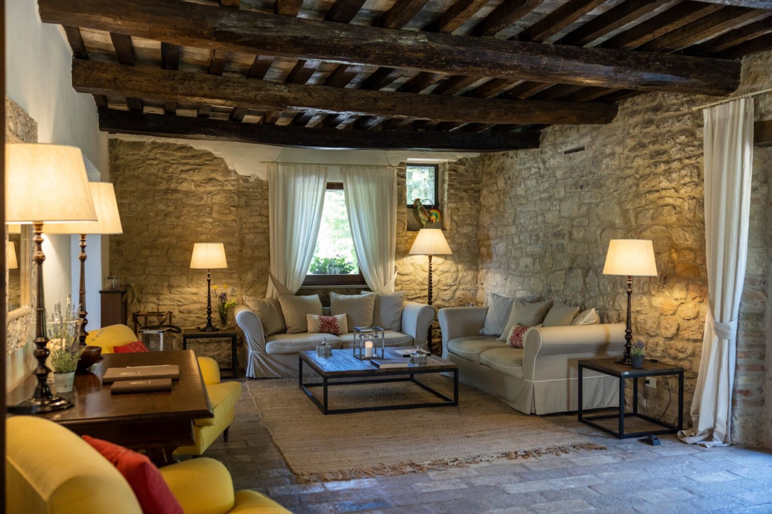 Cozy living room featuring rustic stone walls, wooden beams, and comfortable seating. Natural light filters through sheer curtains, illuminating stylish décor, including a coffee table and elegant lamps. Ideal for relaxation and inviting gatherings. | Only on SopranoVillas