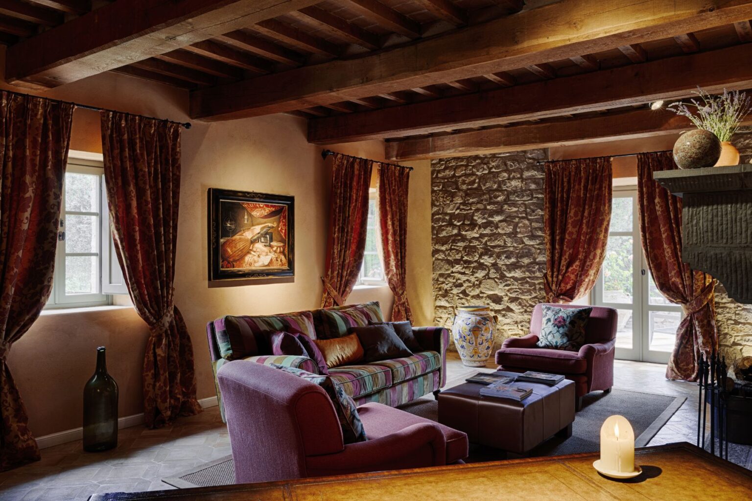 Cozy living room featuring a striped sofa and plush armchairs, adorned with rich curtains and a stone wall. A decorative painting hangs above a stylish coffee table, with a candle providing soft lighting. Large windows allow natural light to fill the space, creating a warm and inviting atmosphere. | Only on SopranoVillas