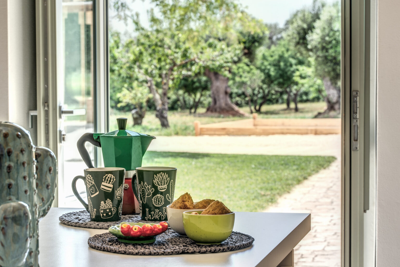 A cozy breakfast setup featuring a green coffee pot, patterned mugs, and a bowl of snacks, with a scenic view of an outdoor garden through an open door. | Only on SopranoVillas
