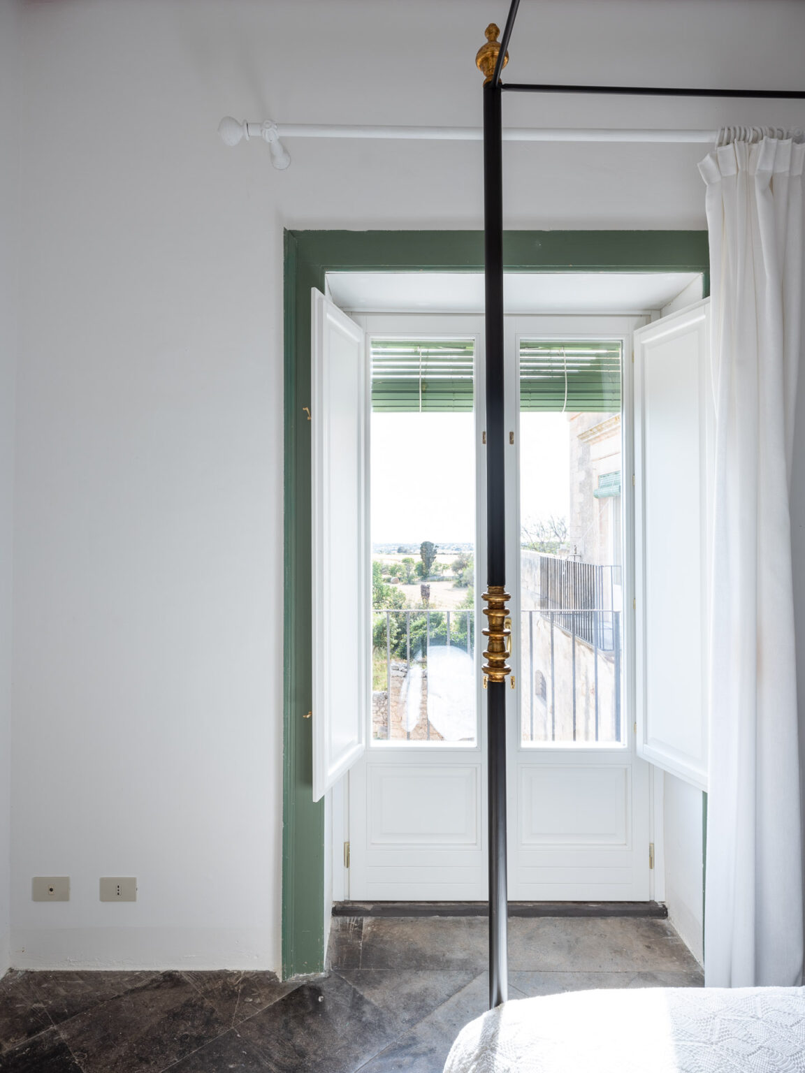 Bright and airy bedroom featuring a large window with open double doors, allowing natural light to flood the space. The window is framed by green trim and offers a view of the outdoor landscape. A stylish black bed frame with a gold accent is positioned in the room, complemented by soft white curtains. The flooring consists of elegant dark marble tiles, enhancing the modern aesthetic. | Only on SopranoVillas