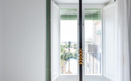Bright and airy bedroom featuring a large window with open double doors, allowing natural light to flood the space. The window is framed by green trim and offers a view of the outdoor landscape. A stylish black bed frame with a gold accent is positioned in the room, complemented by soft white curtains. The flooring consists of elegant dark marble tiles, enhancing the modern aesthetic.