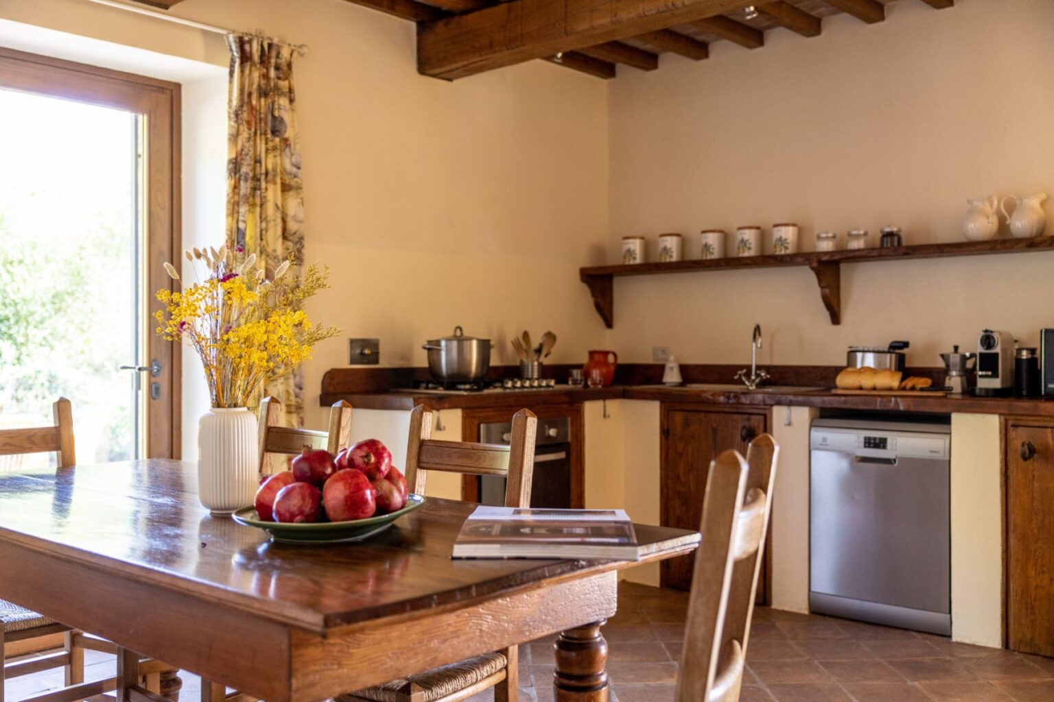 Cozy kitchen and dining area featuring a wooden table with a bowl of red apples, a vase of yellow flowers, and a view of the outdoors through a large window. The kitchen includes modern appliances, wooden cabinets, and a rustic decor style, creating a warm and inviting atmosphere. | Only on SopranoVillas