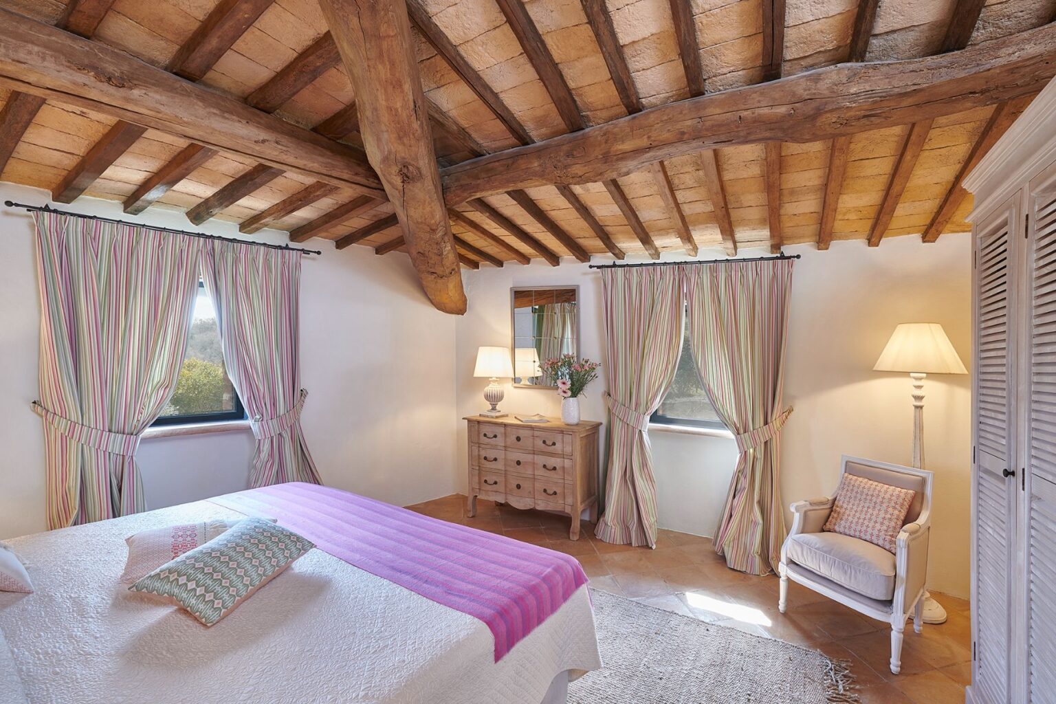 Cozy bedroom featuring rustic wooden beams, striped curtains, and a pastel color scheme. The room includes a comfortable bed with decorative pillows, a wooden dresser, a stylish lamp, and a cozy armchair, creating a warm and inviting atmosphere. | Only on SopranoVillas