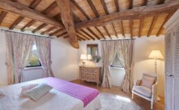 Cozy bedroom featuring rustic wooden beams, striped curtains, and a pastel color scheme. The room includes a comfortable bed with decorative pillows, a wooden dresser, a stylish lamp, and a cozy armchair, creating a warm and inviting atmosphere.