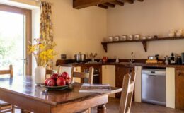 Cozy kitchen and dining area featuring a wooden table with a bowl of red apples, a vase of yellow flowers, and a view of the outdoors through a large window. The kitchen includes modern appliances, wooden cabinets, and a rustic decor style, creating a warm and inviting atmosphere.
