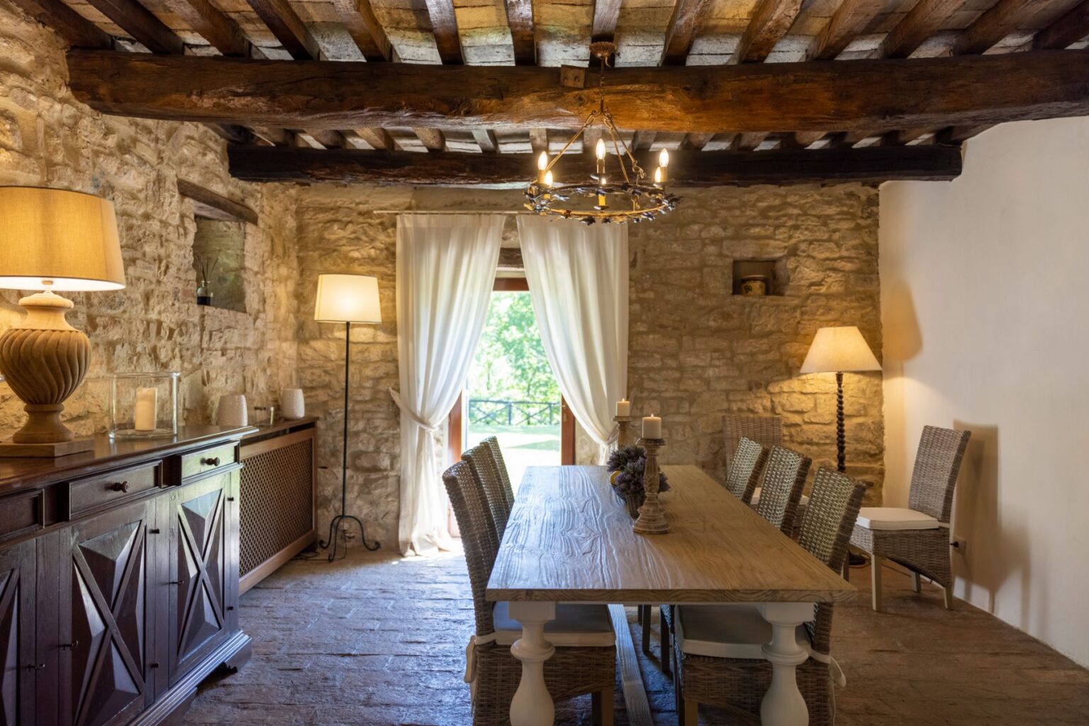 Rustic dining room featuring a long wooden table surrounded by woven chairs, illuminated by a stylish chandelier. The stone walls and wooden beams add a cozy, traditional atmosphere, complemented by soft lighting from lamps and natural light streaming through sheer curtains. | Only on SopranoVillas