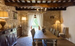 Rustic dining room featuring a long wooden table surrounded by woven chairs, illuminated by a stylish chandelier. The stone walls and wooden beams add a cozy, traditional atmosphere, complemented by soft lighting from lamps and natural light streaming through sheer curtains.