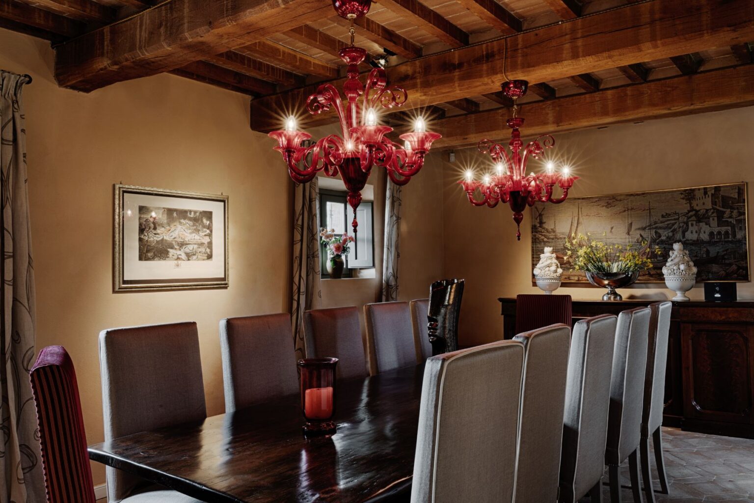 Elegant dining room featuring a long wooden table surrounded by high-backed chairs, adorned with a striking red chandelier and warm lighting. The walls are decorated with artwork and tasteful floral arrangements, creating a cozy and inviting atmosphere. | Only on SopranoVillas