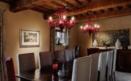 Elegant dining room featuring a long wooden table surrounded by high-backed chairs, adorned with a striking red chandelier and warm lighting. The walls are decorated with artwork and tasteful floral arrangements, creating a cozy and inviting atmosphere.