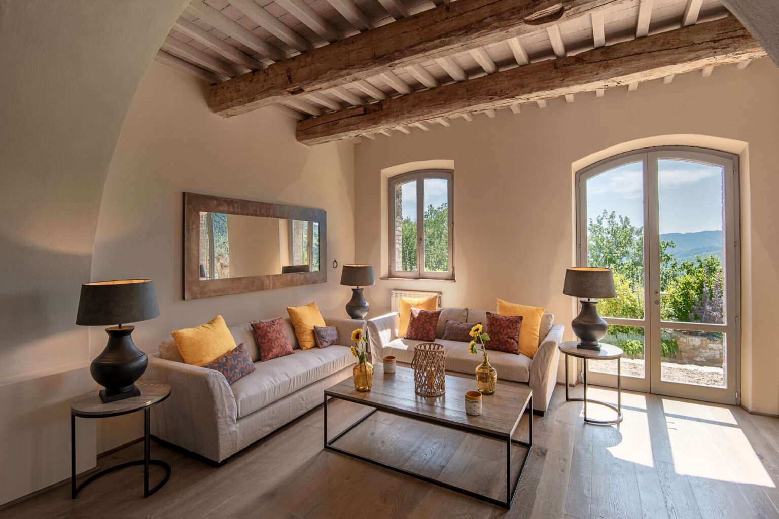 Bright and airy living room with rustic wooden beams, featuring a cozy beige sofa adorned with colorful cushions, a glass coffee table, and decorative vases. Large windows offer scenic views of greenery, enhancing the inviting atmosphere. Ideal for relaxation and stylish interior inspiration. | Only on SopranoVillas
