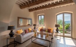Bright and airy living room with rustic wooden beams, featuring a cozy beige sofa adorned with colorful cushions, a glass coffee table, and decorative vases. Large windows offer scenic views of greenery, enhancing the inviting atmosphere. Ideal for relaxation and stylish interior inspiration.