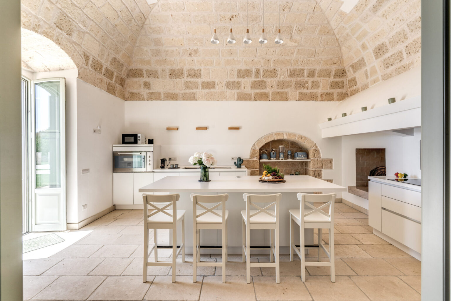 Spacious modern kitchen with high vaulted ceilings, featuring a large white island, wooden bar stools, and natural stone walls. Brightly lit with large windows, the kitchen includes modern appliances and a cozy fireplace, creating an inviting atmosphere. | Only on SopranoVillas