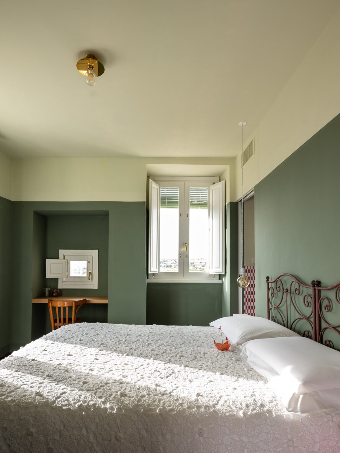 Cozy bedroom with a double bed featuring a white textured quilt, complemented by a decorative red metal headboard. Natural light streams in through open double windows, illuminating a small desk with a wooden chair and a decorative item. The room is painted in soothing green tones, creating a tranquil atmosphere. | Only on SopranoVillas