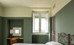 Cozy bedroom with a double bed featuring a white textured quilt, complemented by a decorative red metal headboard. Natural light streams in through open double windows, illuminating a small desk with a wooden chair and a decorative item. The room is painted in soothing green tones, creating a tranquil atmosphere.