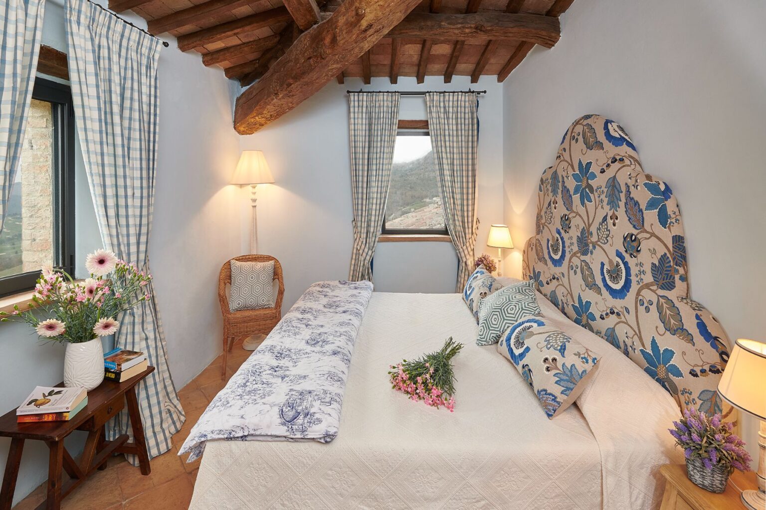 Cozy and elegantly decorated bedroom featuring a king-size bed with floral-patterned pillows, wooden beams, and large windows with checkered curtains. A bedside table holds books and a vase of flowers, adding a touch of charm to the inviting space. | Only on SopranoVillas