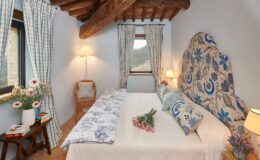 Cozy and elegantly decorated bedroom featuring a king-size bed with floral-patterned pillows, wooden beams, and large windows with checkered curtains. A bedside table holds books and a vase of flowers, adding a touch of charm to the inviting space.